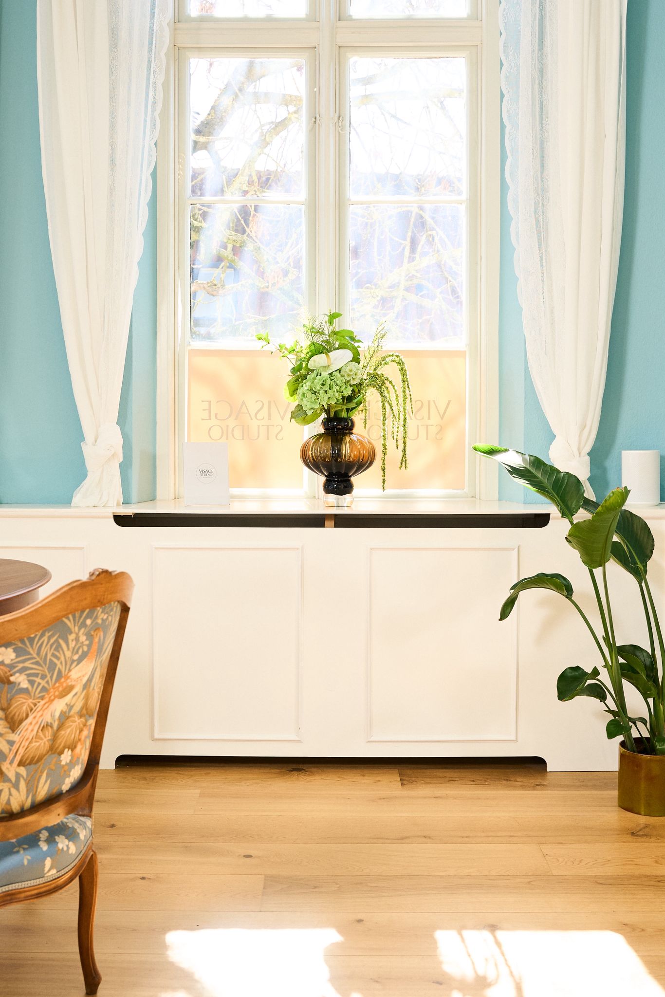 Light-filled interior at Visage Studio featuring original sash windows, soft blue walls, decorative moulding, and fresh floral styling.
